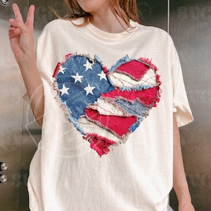 May include: Cream-coloured t-shirt with a heart-shaped design made from denim and red fabric patches. The denim section features white stars, resembling the American flag. The edges of the patches are frayed.