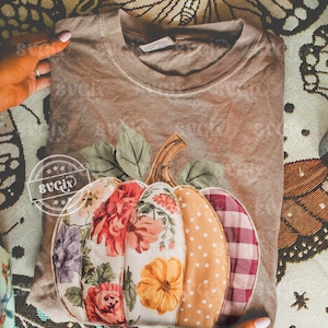 May include: A brown t-shirt featuring a decorative pumpkin design. The pumpkin is made of fabric with floral, polka dot, and plaid patterns in shades of orange, red, and purple. Green leaves and a brown stem complete the design.