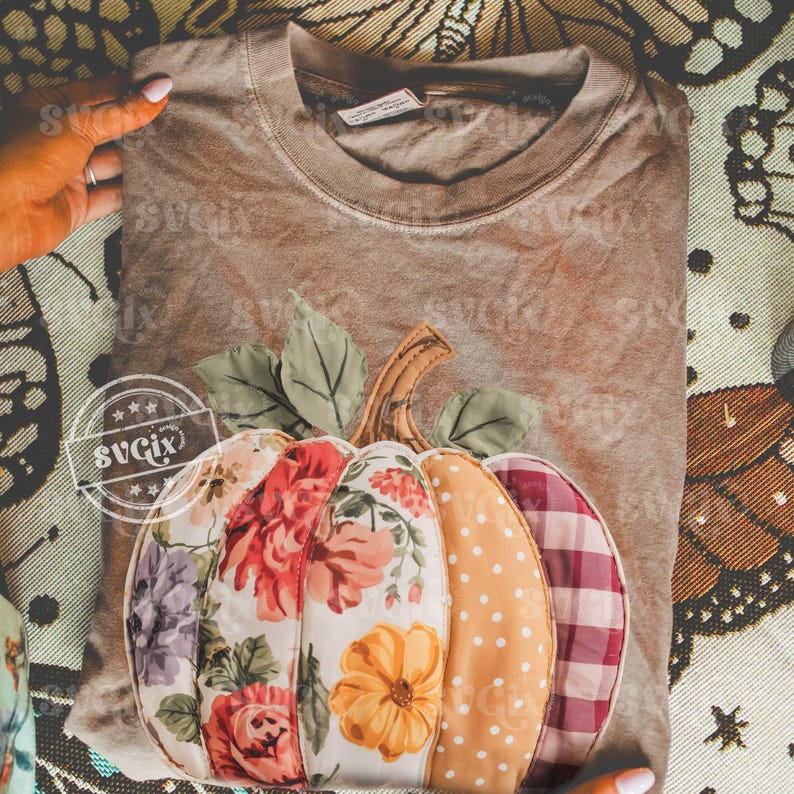 May include: A brown t-shirt featuring a decorative pumpkin design. The pumpkin is made of fabric with floral, polka dot, and plaid patterns in shades of orange, red, and purple. Green leaves and a brown stem complete the design.
