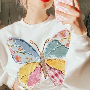 May include: White sweatshirt featuring a colorful butterfly applique. The butterfly design incorporates various fabrics in shades of blue, pink, yellow, and floral patterns. The sweatshirt has long sleeves and a crew neck.