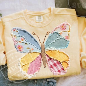 May include: A light yellow sweatshirt with a large, colorful butterfly applique. The butterfly wings are made of patchwork fabric in shades of blue, pink, yellow, and green, with floral and gingham patterns. The sweatshirt has a crew neck and long sleeves.