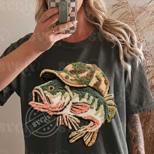 May include: Dark green t-shirt with an embroidered design of a fish wearing a camouflage hat. The fish is detailed with green, orange, and beige colours. The design is centrally placed on the shirt, creating a unique graphic.