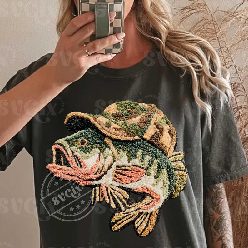 May include: Dark green t-shirt with an embroidered design of a fish wearing a camouflage hat. The fish is detailed with green, orange, and beige colours. The design is centrally placed on the shirt, creating a unique graphic.