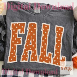 May include: A dark gray t-shirt with the word "FALL" in large, orange, polka-dotted letters. The letters have a white outline. The shirt is folded, and a plaid pattern is visible at the bottom.