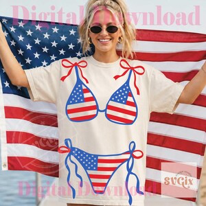 May include: Cream-colored t-shirt featuring a bikini design with the American flag. The bikini top has red bows, and the bottom has blue ties. The background includes an American flag.