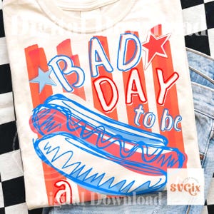 May include: Cream-colored t-shirt with a graphic design featuring the text "BAD DAY to be" in red and blue, along with a cartoon hot dog. The design includes red and white stripes and star accents.