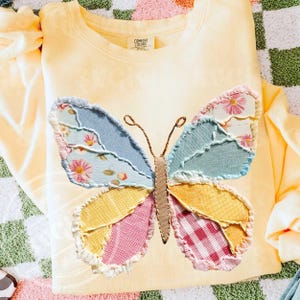May include: A light yellow Comfort Colors sweatshirt featuring a large, patchwork butterfly applique. The butterfly wings are made of various patterned fabrics in blue, pink, yellow, and floral prints. The sweatshirt has a crew neck and long sleeves.