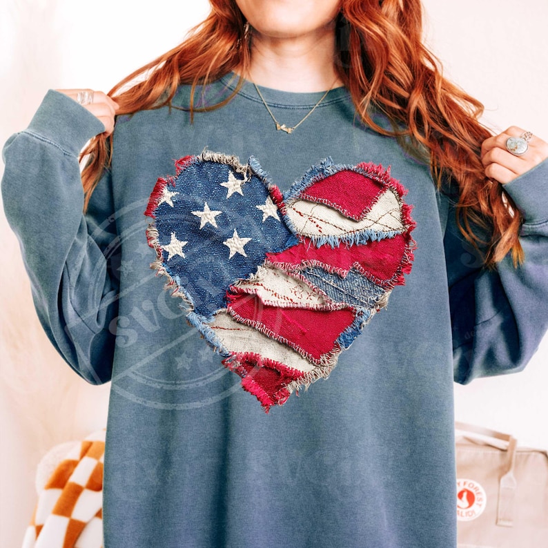 May include: A blue-grey sweatshirt with a heart-shaped design made from patches. The heart design incorporates elements of the American flag, with blue denim and white stars on one side, and red fabric on the other. The sweatshirt has long sleeves.