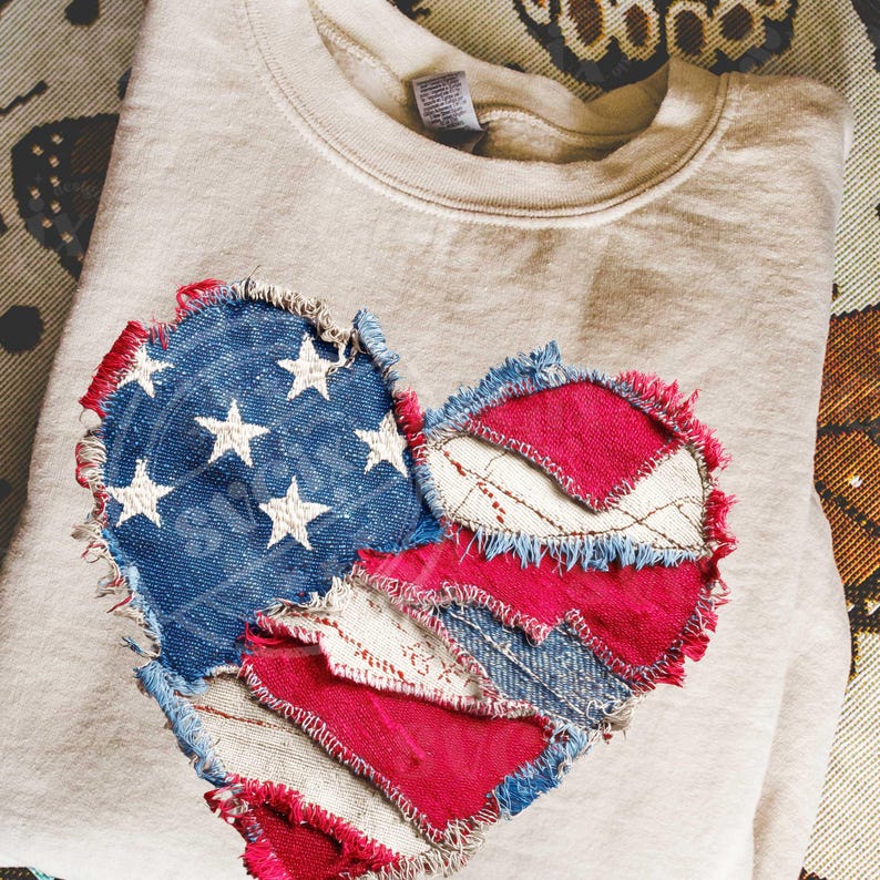 May include: A beige sweatshirt featuring a heart-shaped patch. The patch is crafted from denim and red fabric, with white stars on a blue denim section. The edges are frayed, creating a distressed appearance. The sweatshirt is folded.