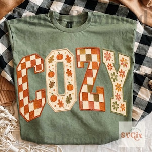 May include: A sage green t-shirt featuring the word COZY in large block letters. The C and Z have a checkered pattern in orange and white, the O contains orange pumpkins and autumn leaves, and the Y features orange and green flower motifs.
