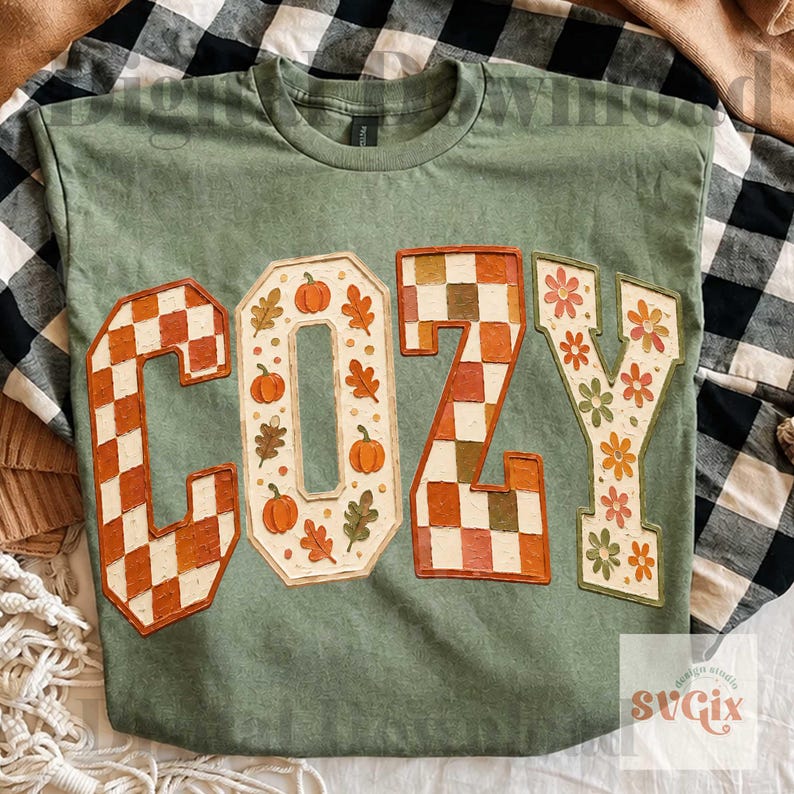 May include: A sage green t-shirt featuring the word COZY in large block letters. The C and Z have a checkered pattern in orange and white, the O contains orange pumpkins and autumn leaves, and the Y features orange and green flower motifs.