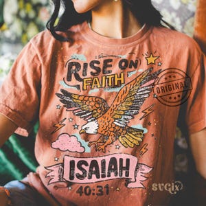 Puede incluir: Camiseta de color coral con un dise&ntilde;o de un &aacute;guila volando. El dise&ntilde;o incluye las palabras "RISE ON FAITH" sobre el &aacute;guila e "ISAIAH 40:31" debajo. La palabra "ORIGINAL" est&aacute; estampada en el lado derecho.