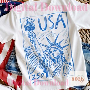 K&ouml;nnte beinhalten: Wei&szlig;es T-Shirt mit einer blauen Skizze der Freiheitsstatue, dem Wort "USA" und der Zahl "250". Das Design befindet sich in einem rechteckigen Rahmen. Das Shirt befindet sich auf einem Hintergrund mit einer amerikanischen Flagge und Denim.