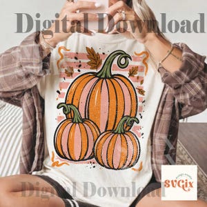 May include: Cream-colored t-shirt featuring a graphic of three pumpkins in shades of orange and pink, with green stems and autumn leaves. The design is set against a pink striped background. The words "Digital Download" are visible.