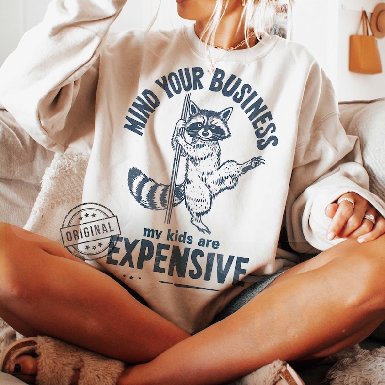 Pu&ograve; includere: Felpa color crema con grafica di procione blu navy che tiene un palo, e il testo "MIND YOUR BUSINESS" e "my kids are EXPENSIVE". La parola "ORIGINAL" &egrave; stampata sul lato sinistro.