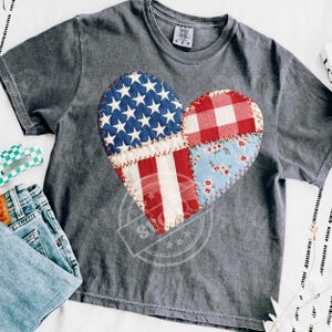 May include: Gray t-shirt with a heart-shaped patchwork design. The heart features sections with the American flag, red and white gingham, and a floral pattern. The shirt is a Comfort Colors brand.