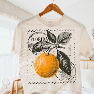 May include: Off-white t-shirt with a retro design of an orange, leaves, and the word "FLORIDA" within a circular stamp. The graphic is in orange, black, and green. The t-shirt is hanging on a wooden hanger.