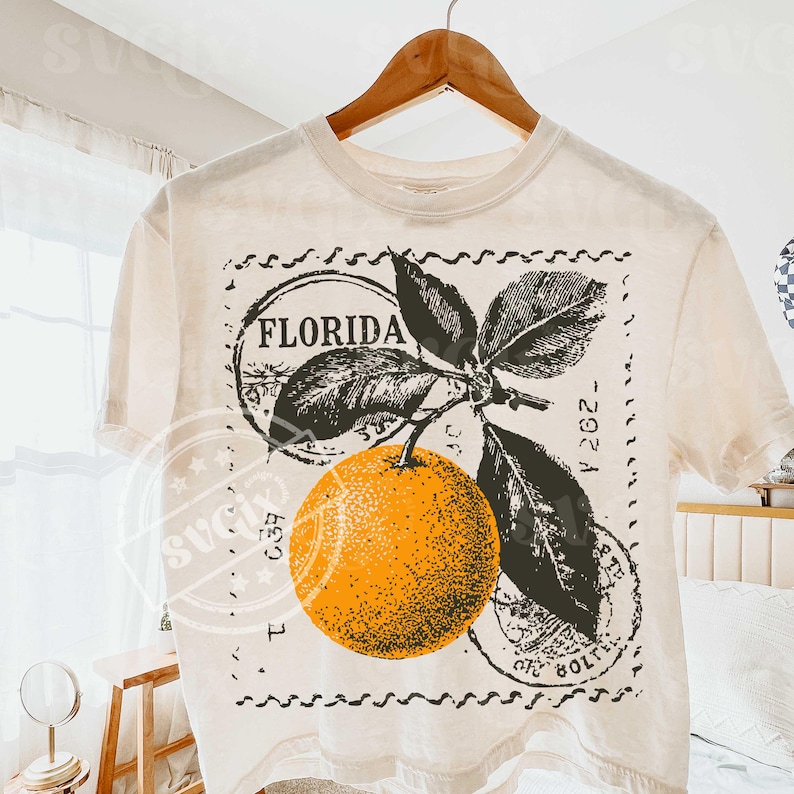 May include: Off-white t-shirt with a retro design of an orange, leaves, and the word "FLORIDA" within a circular stamp. The graphic is in orange, black, and green. The t-shirt is hanging on a wooden hanger.