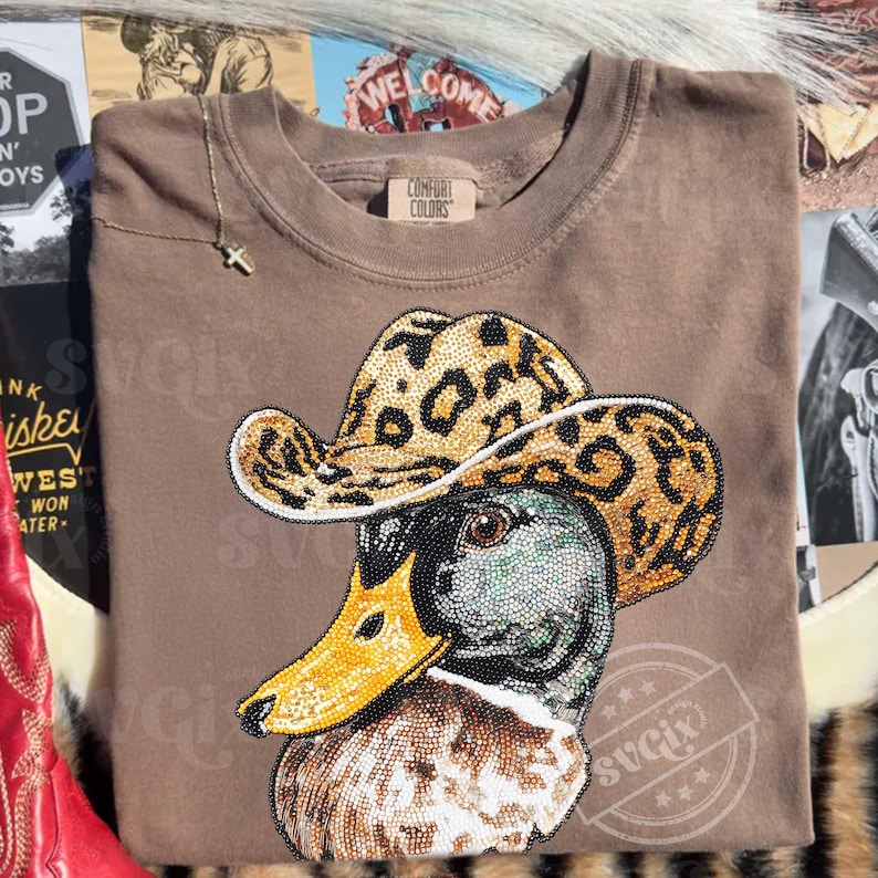 May include: Brown Comfort Colors sweatshirt featuring a detailed graphic of a duck wearing a leopard print cowboy hat. The duck's head is embellished with small, sparkling beads. A gold necklace with a cross hangs on the shirt.