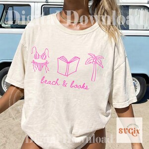 K&ouml;nnte beinhalten: Cremefarbenes T-Shirt mit einer rosa Grafik eines Bikinis, eines offenen Buches und einer Palme. Die Worte "beach & books" sind in rosa Schrift darunter geschrieben. Das Shirt hat kurze &Auml;rmel.