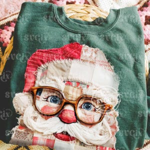 May include: Green sweatshirt featuring a patchwork Santa Claus face with glasses. The Santa design includes a red hat, white beard, and blue eyes. The sweatshirt is folded on a woven basket, with a floral patterned blanket in the background.