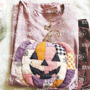 May include: A light purple t-shirt with a patchwork pumpkin design. The pumpkin is made of various fabrics in shades of orange, pink, purple, and black, with a smiling jack-o'-lantern face. A gold butterfly necklace rests on the shirt.