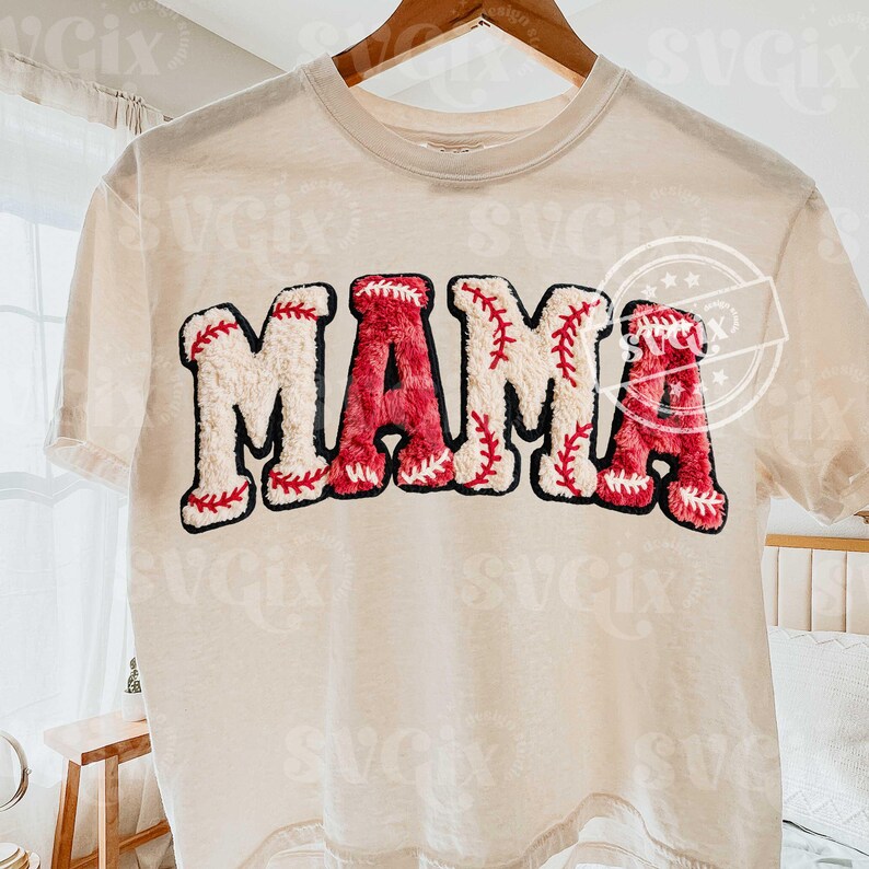 May include: Cream-colored t-shirt with the word "MAMA" in a baseball-themed design. The letters are white with red baseball stitching and a red baseball texture. The shirt is hanging on a wooden hanger.