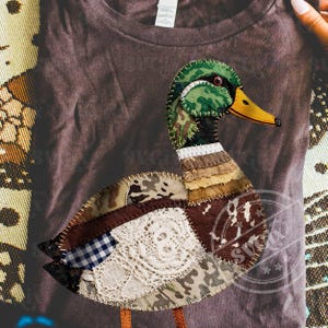 May include: A brown t-shirt featuring a patchwork duck design. The duck has a green and camouflage head, a yellow beak, and a body composed of various fabric patterns, including lace and plaid. The duck's legs are brown.