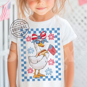 May include: A white t-shirt with a graphic of a goose wearing a red, white, and blue bandana and sunglasses, holding a hot dog and an American flag. The goose is standing on a blue and white checkered background with red, white, and blue flowers.
