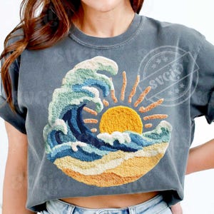 May include: A grey t-shirt with an embroidered design of a wave, sun, and beach. The wave is in blue and green, with a yellow sun and sandy beach. The shirt has short sleeves. The design is circular.