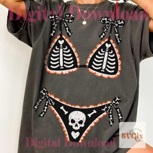 Peut inclure: Un t-shirt gris anthracite arborant un imprim&eacute; graphique repr&eacute;sentant un bikini noir avec des d&eacute;tails de cage thoracique squelettique blanche sur le haut et un motif de cr&acirc;ne sur le bas, agr&eacute;ment&eacute; de bords &agrave; volants orange et de n&oelig;uds &agrave; motifs d'os.