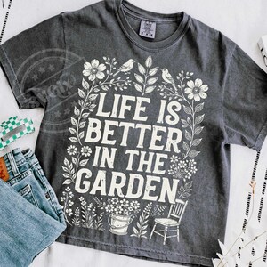 Puede incluir: Camiseta gris oscuro con el texto blanco "LIFE IS BETTER IN THE GARDEN". El dise&ntilde;o incluye elementos florales, p&aacute;jaros, un cubo y una silla. La camiseta se muestra con vaqueros azules.