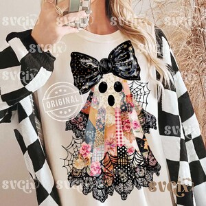 May include: Cream-colored Halloween-themed t-shirt featuring a patchwork ghost design with a black bow, lace trim, and spiderwebs. The ghost's body is made of floral and patterned fabrics. The word "ORIGINAL" is stamped on the shirt.