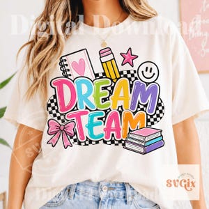 Pode incluir: Camiseta creme com um gráfico colorido "DREAM TEAM". O design inclui um caderno, lápis, estrela, carinha sorridente, laço e livros. As letras são multicoloridas em um fundo quadriculado preto e branco.