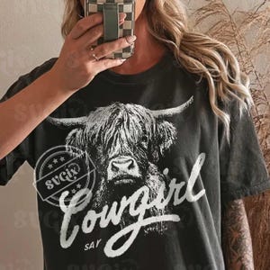 May include: Dark gray t-shirt featuring a black and white illustration of a Highland cow. The shirt has the word "Cowgirl" in a white script font, with the word "Say" below it. A circular logo with stars is also present.