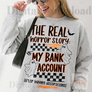 Pode incluir: Camisola cinzenta com o texto "THE REAL horror story: MY BANK ACCOUNT after buying decorations!" em preto e laranja. O design inclui um morcego, duas ilustra&ccedil;&otilde;es de fantasmas e um padr&atilde;o xadrez, ideal para o Halloween.