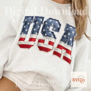 May include: A light gray sweatshirt with the word "USA" appliqued on the front. The letters are made of denim fabric with a blue star pattern and red and white stripes, with a frayed edge.