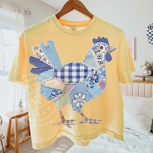 May include: Yellow t-shirt with a patchwork rooster design. The rooster features blue, white, and floral patterned fabric. The shirt is hanging on a wooden hanger.