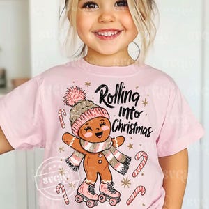 May include: Pale pink t-shirt with a gingerbread person design. The gingerbread figure wears a hat, scarf, and roller skates, with the words "Rolling Into Christmas." Candy canes and stars complete the festive design.