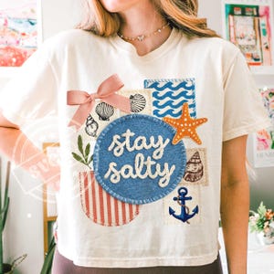 Peut inclure: T-shirt cr&egrave;me avec un motif nautique. Il pr&eacute;sente un cercle bleu avec le texte "stay salty", un n&oelig;ud rose, des coquillages, une &eacute;toile de mer et une ancre. Le motif comprend des rayures rouges et blanches et des motifs de vagues bleues.