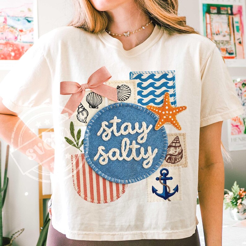 Peut inclure: T-shirt cr&egrave;me avec un motif nautique. Il pr&eacute;sente un cercle bleu avec le texte "stay salty", un n&oelig;ud rose, des coquillages, une &eacute;toile de mer et une ancre. Le motif comprend des rayures rouges et blanches et des motifs de vagues bleues.