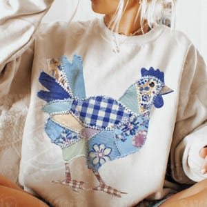 May include: Cream-colored sweatshirt featuring a patchwork rooster design. The rooster is composed of various blue fabrics, including gingham, floral, and solid patterns. The rooster's legs are a brown and white checkered pattern.