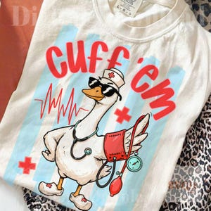 Op de afbeelding: Crèmekleurig T-shirt met een cartoon gans in een verpleegstersoutfit, met een zonnebril en een stethoscoop. De tekst "Cuff 'em" is rood. Het ontwerp bevat een hartslaglijn en medische kruizen.