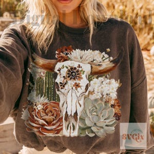 Puede incluir: Una sudadera de cuello redondo color marr&oacute;n oscuro con un estampado gr&aacute;fico de un cr&aacute;neo de longhorn blanco adornado con varias flores, incluyendo flores blancas, detalles florales marrones, cactus verdes y una suculenta verde grande.