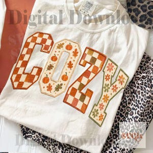 May include: A white t-shirt featuring the word COZY in large block letters. The C and Z are filled with an orange and white checkerboard pattern. The O is decorated with orange pumpkins and leaves, and the Y features orange and green flowers.