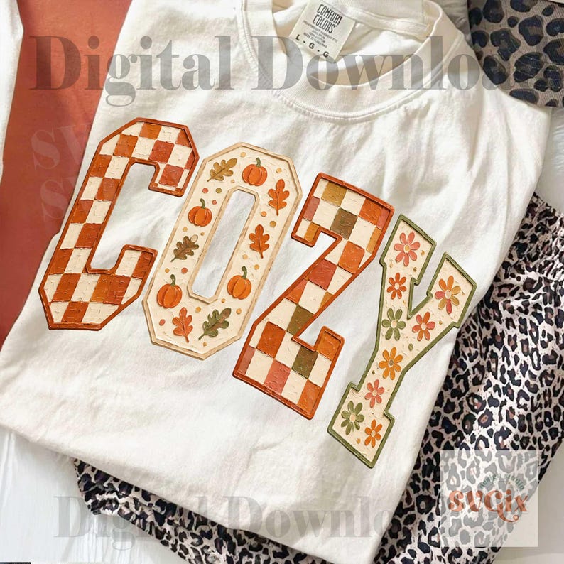 May include: A white t-shirt featuring the word COZY in large block letters. The C and Z are filled with an orange and white checkerboard pattern. The O is decorated with orange pumpkins and leaves, and the Y features orange and green flowers.