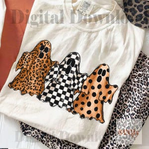 May include: A cream-colored t-shirt featuring a graphic of three ghosts. The left ghost has a brown leopard print, the middle ghost has a black and white checkerboard pattern, and the right ghost has a brown polka dot pattern on a tan background.