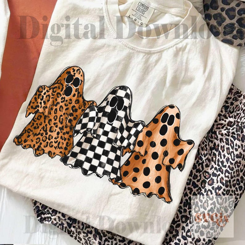 May include: A cream-colored t-shirt featuring a graphic of three ghosts. The left ghost has a brown leopard print, the middle ghost has a black and white checkerboard pattern, and the right ghost has a brown polka dot pattern on a tan background.