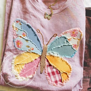 May include: A light pink t-shirt with a large, colorful butterfly applique. The butterfly wings are made of patchwork fabric in shades of blue, yellow, pink, and red. A gold butterfly pendant necklace is near the neckline. The shirt is casual and handmade.
