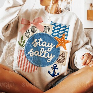 Peut inclure: Sweat-shirt cr&egrave;me avec un motif patchwork. Il pr&eacute;sente un n&oelig;ud rose, des coquillages, une &eacute;toile de mer et l'expression "stay salty" en &eacute;criture blanche sur un cercle bleu. D'autres patchs incluent une ancre et un motif de vagues.
